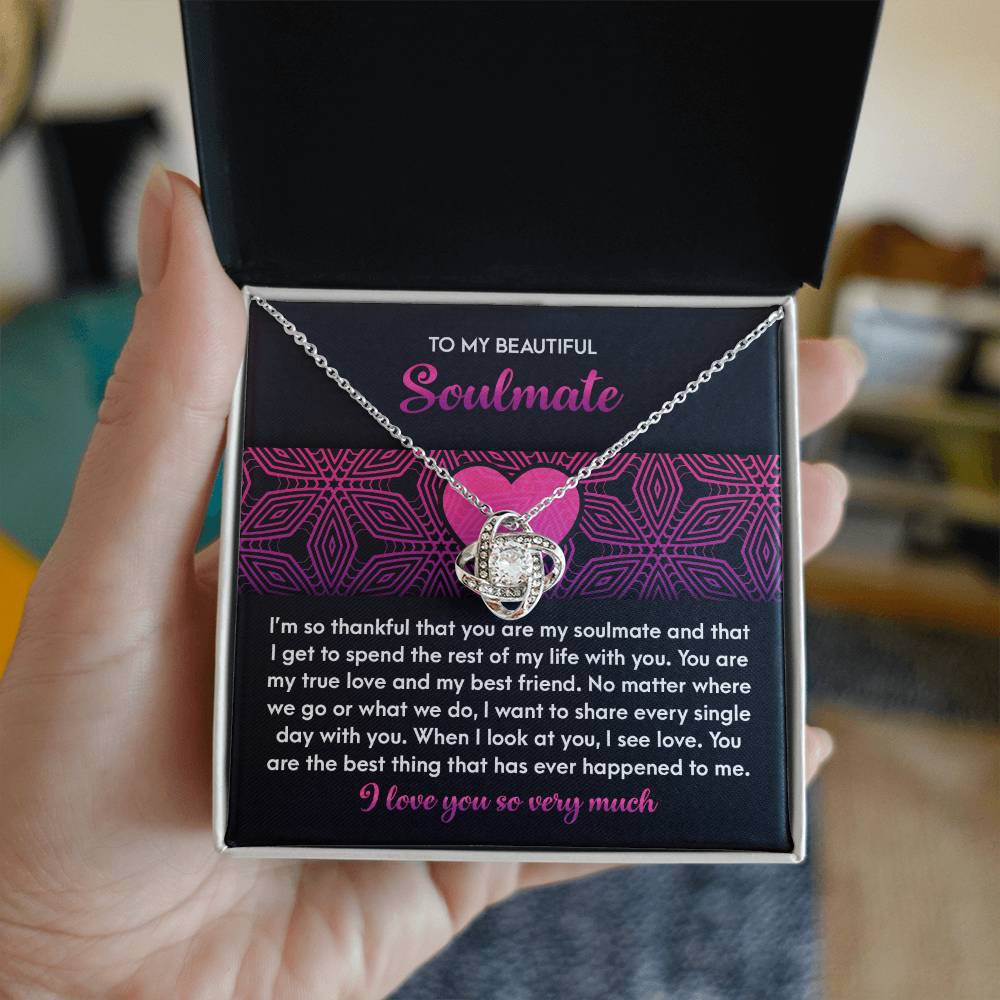 A hand gently holds a Soulmate-I See Love - Love Knot Necklace in a black gift box, featuring a silver pendant with cubic zirconia crystals. A pink and purple card accompanies it, conveying a heartfelt message about the unbreakable bond with your soulmate.