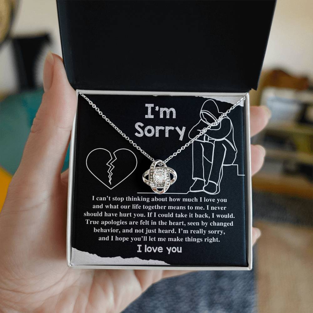 The "Sorry-Not Just Heard - Love Knot Necklace" features an apology message on a black background. The heartfelt text expresses regret and a promise to change, accompanied by illustrations of a heart and a person sitting with their head down. Cubic zirconia crystals add a touch of elegance, with "I love you" written at the bottom.