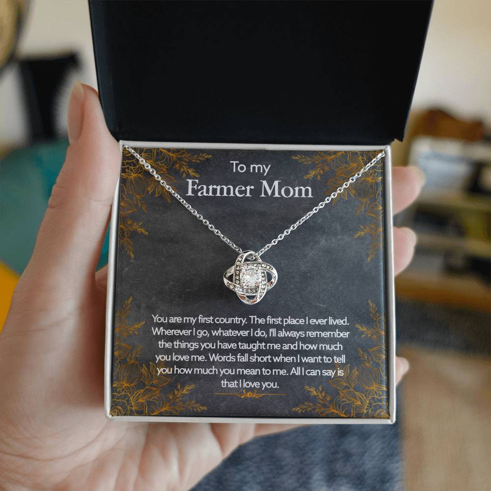 A hand is holding an open jewelry box with a To My Farmer Mom, You Are My First Country - Love Knot Necklace and pendant, adorned with cubic zirconia crystals, inscribed with a heartfelt message dedicated to a "farmer mom.