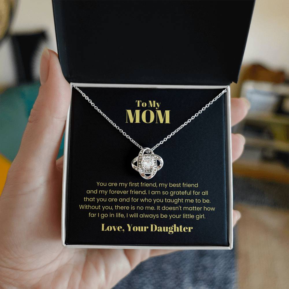 A hand holds an open box displaying the stunning "To My Mom, You Are My First Friend - Love Knot Necklace," adorned with cubic zirconia crystals. The box contains a heartfelt message to "Mom" from a daughter, expressing gratitude and love.