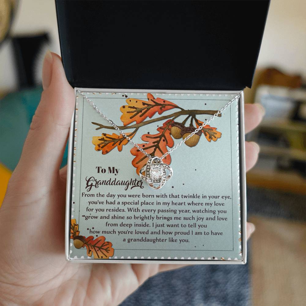 A hand holds an open box showcasing the "Granddaughter-From Deep Inside - Love Knot Necklace," featuring a heart-shaped pendant embellished with cubic zirconia crystals. The box includes a decorative background and a heartfelt message tailored for a granddaughter, creating an ideal personalized gift.