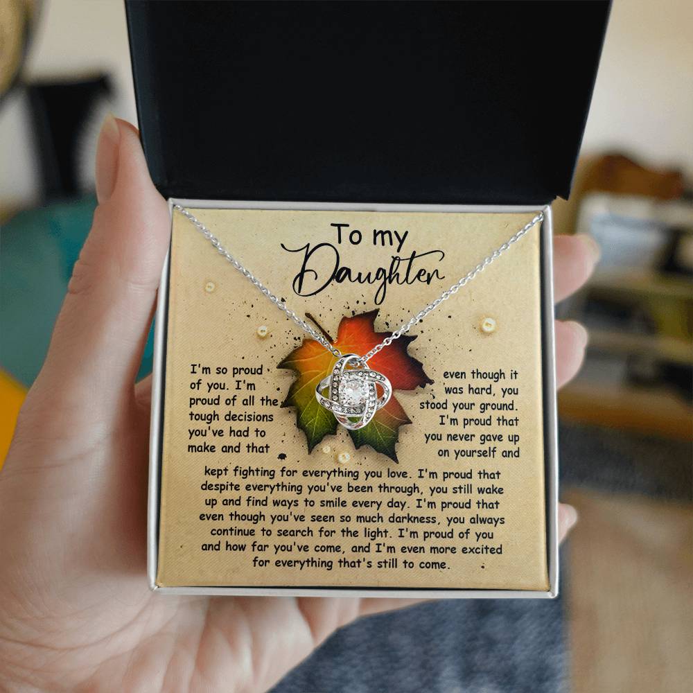A hand holds an open box with a Daughter-Stood Your Ground - Love Knot Necklace, adorned with cubic zirconia crystals and accompanied by a heartfelt message to a daughter, expressing pride, acknowledgment of struggles, and encouragement for the future.