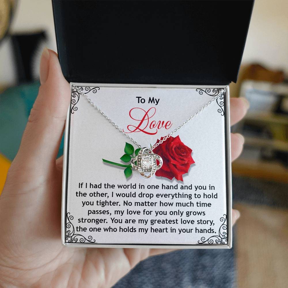 A hand holds an open black box revealing the Soulmate-In Your Hands - Love Knot Necklace adorned with sparkling cubic zirconia. The lid features a note addressed "To My Love," with a red rose and a heartfelt message, creating the ideal personalized gift.