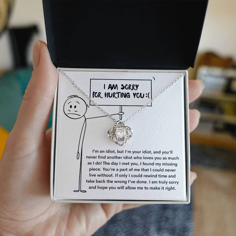 A hand holds an open box displaying the "Sorry-Missing Piece - Love Knot Necklace" adorned with cubic zirconia crystals on a card that says "I AM SORRY FOR HURTING YOU!" accompanied by an apology message and a drawing of a stick figure holding a heart.