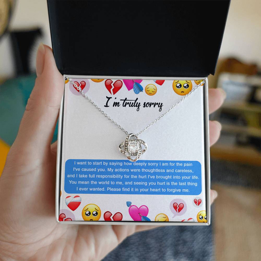 The Sorry-How Deeply Sorry - Love Knot Necklace features a pendant with an intertwined design, adorned with sparkling cubic zirconia crystals. The piece rests on a card that includes an apology message accompanied by various emoticons, such as hearts and sad faces.