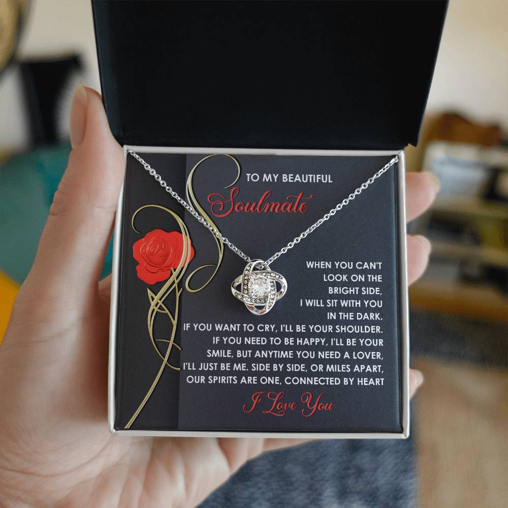 A hand holds a Soulmate-Be Me - Love Knot Necklace embellished with cubic zirconia crystals, next to a card featuring a red rose and a touching message about being there for your soulmate.