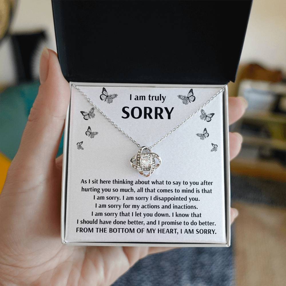 A hand holds a jewelry box containing the Sorry-Done Better - Love Knot Necklace, crafted from 14k white gold. The box lid is adorned with the message "I am truly SORRY," encircled by butterflies. Below it, a heartfelt apology note vows improvement and remorse, complemented by the premium cubic zirconia embellishments on the necklace.