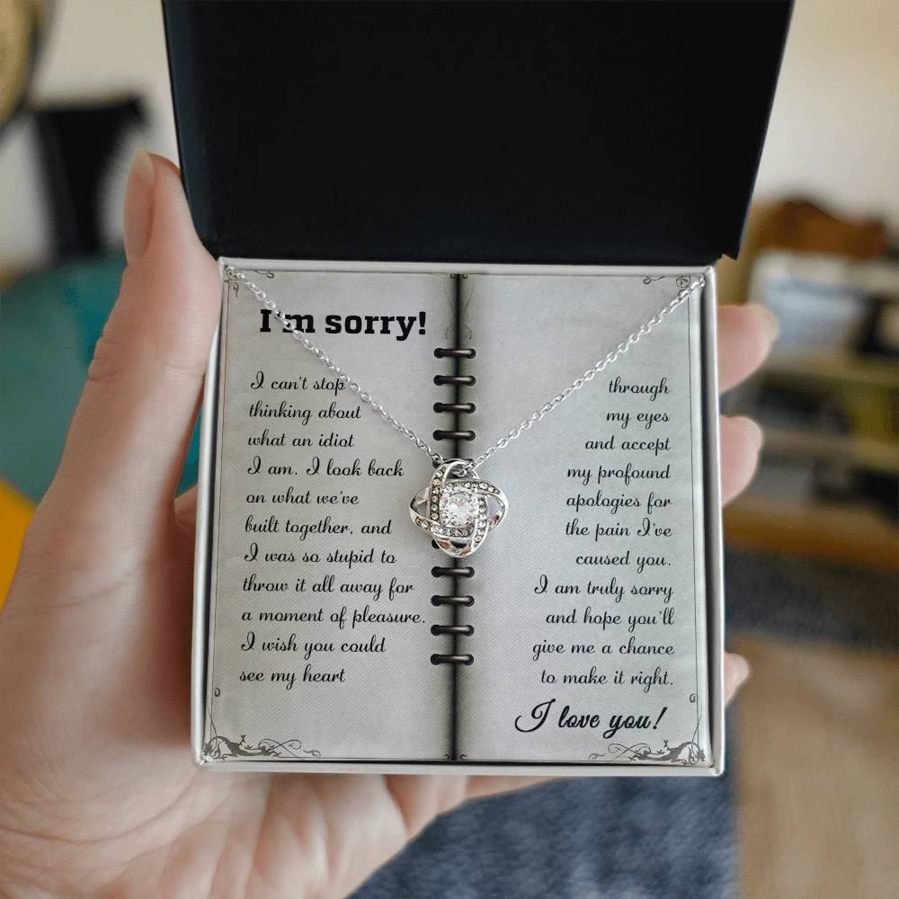A hand holds an open gift box containing a stunning Sorry-What An Idiot - Love Knot Necklace adorned with premium cubic zirconia crystals and a note that reads, "I'm sorry! I can't stop thinking about what an idiot I am. I was so wrong about what we built...I love you.