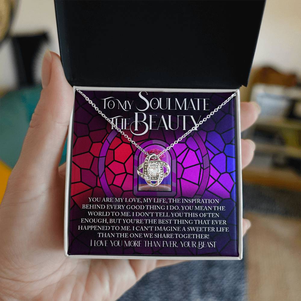 A hand holds an open box revealing the Soulmate-Sweeter Life Love Knot Necklace adorned with cubic zirconia crystals. The backdrop features a purple and pink stained glass design with a romantic message, enhancing this elegant piece's allure.