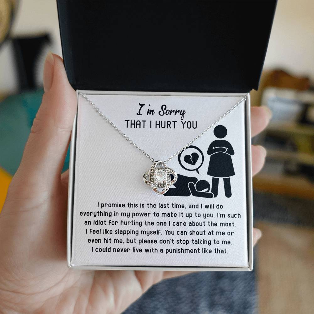 A hand holding an open gift box with a Sorry-Stop Talking - Love Knot Necklace adorned with Cubic Zirconia Crystals. Inside the box, there's an apology message featuring a stick figure illustration of a heartbroken scene, along with text expressing regret and a promise to make amends.