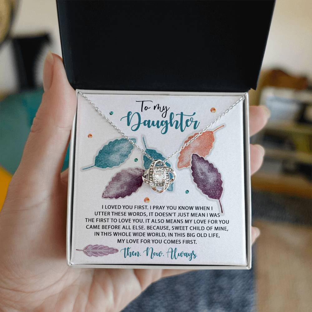The "Daughter-Pray You Know - Love Knot Necklace" features a heart-shaped pendant adorned with cubic zirconia crystals, and comes in a gift box decorated with colorful leaf designs and a heartfelt message addressed to a daughter. The classic gift is beautifully presented in an open box cradled by a hand.