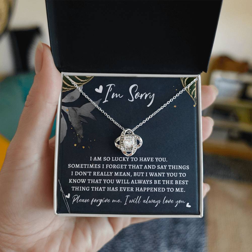 A person holds an open box containing a "Sorry-The Best Thing - Love Knot Necklace" and an apology message, reading, "I’m Sorry. I am so lucky to have you... Please forgive me. I will always love you." This personalized gift with its premium cubic zirconia sparkles as a testament to their heartfelt emotions.
