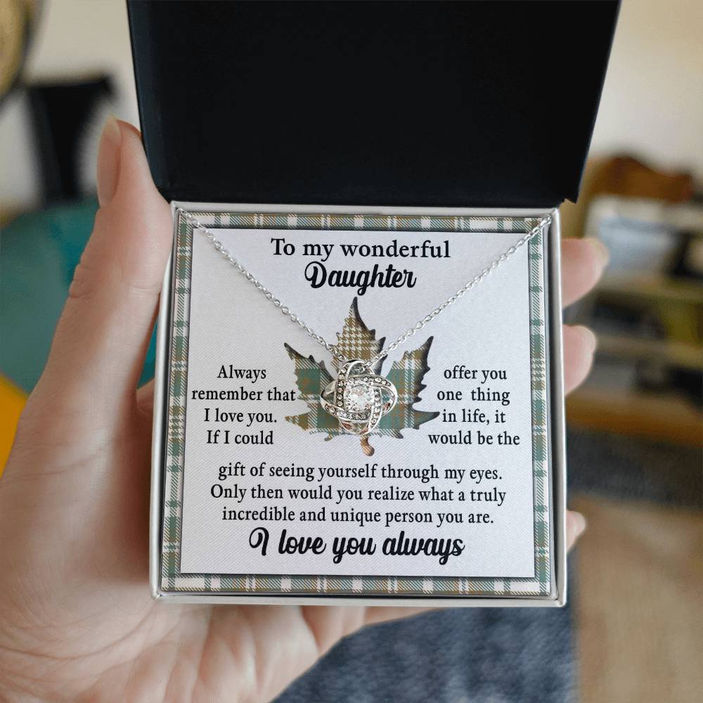 A hand holding an open jewelry box containing the Daughter-Through My Eyes - Love Knot Necklace, featuring a unique pendant adorned with cubic zirconia crystals and a gold finish, accompanied by a heartfelt message addressed to a daughter, expressing love and appreciation.