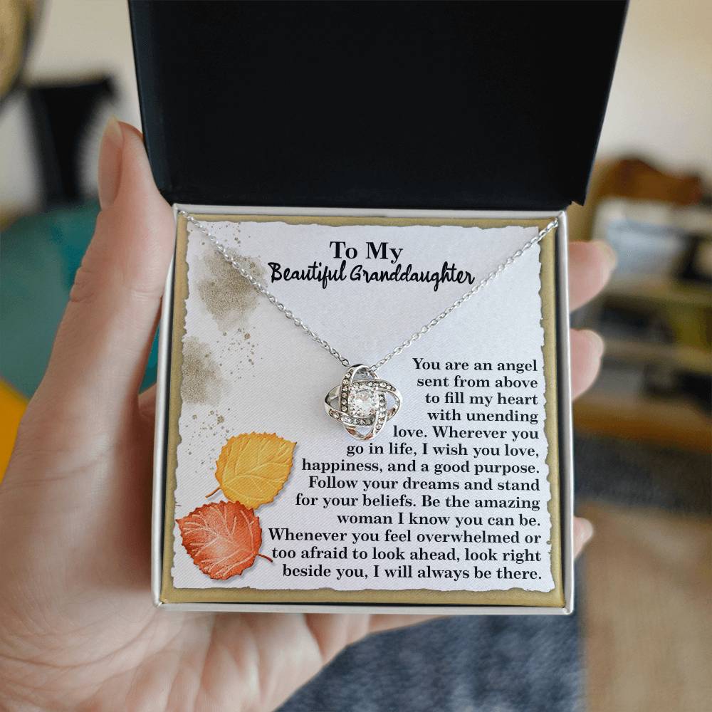 A Granddaughter-An Angel Love Knot Necklace, embellished with cubic zirconia crystals, is beautifully presented in an open box accompanied by a heartfelt message for a granddaughter. The backdrop showcases autumn leaves and elegant text, making it the perfect personalized gift.