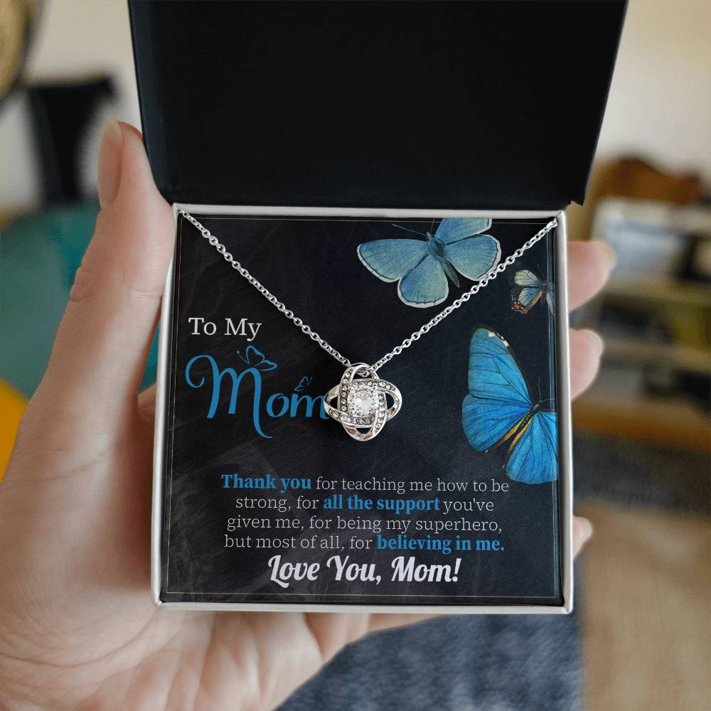 A hand holds an open box containing a "To Mom, Believing In Me - Love Knot Necklace" adorned with cubic zirconia crystals and a card that reads, "To My Mom," with a thank you message, decorated with images of blue butterflies.