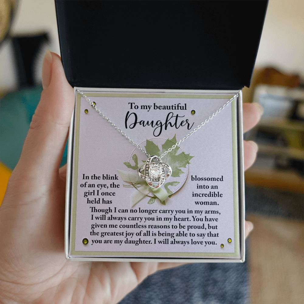 A person holds an open jewelry box containing a Daughter-Incredible Woman Love Knot Necklace adorned with cubic zirconia crystals and a heartfelt note addressed to their daughter, expressing love and pride.