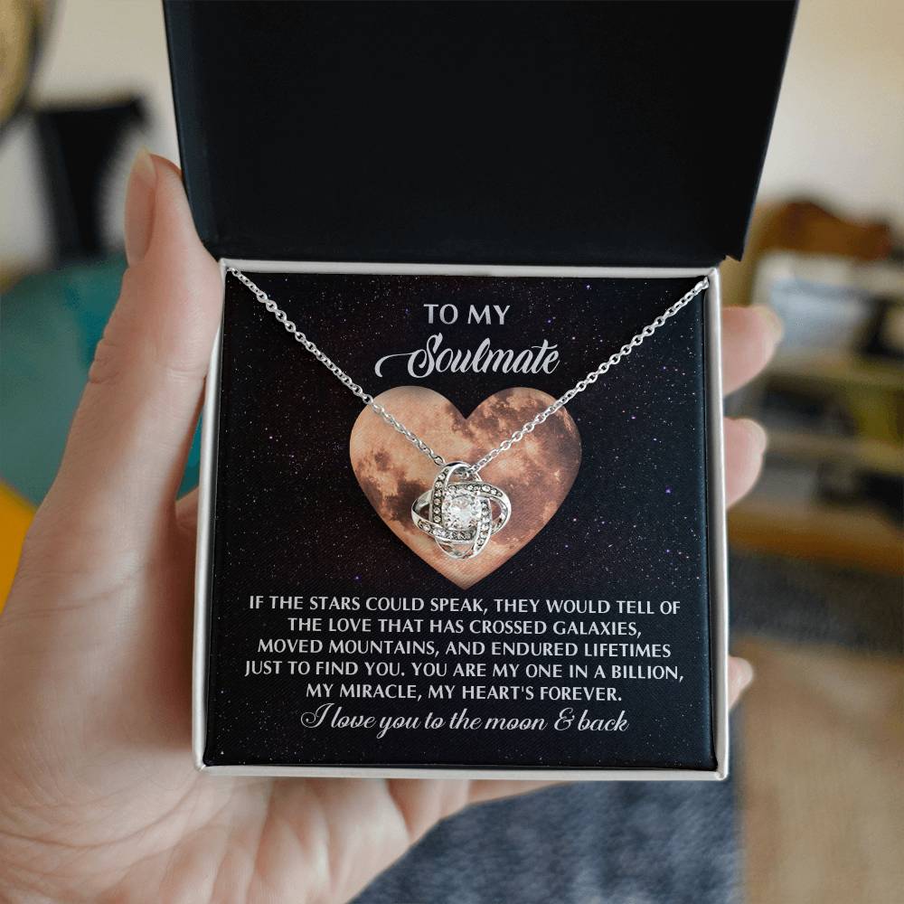 A hand delicately holds an open jewelry box revealing the stunning Soulmate-Crossed Galaxies Love Knot Necklace, adorned with cubic zirconia crystals. The box has a heart-shaped image and a heartfelt love and soulmate message, making it the perfect personalized gift.