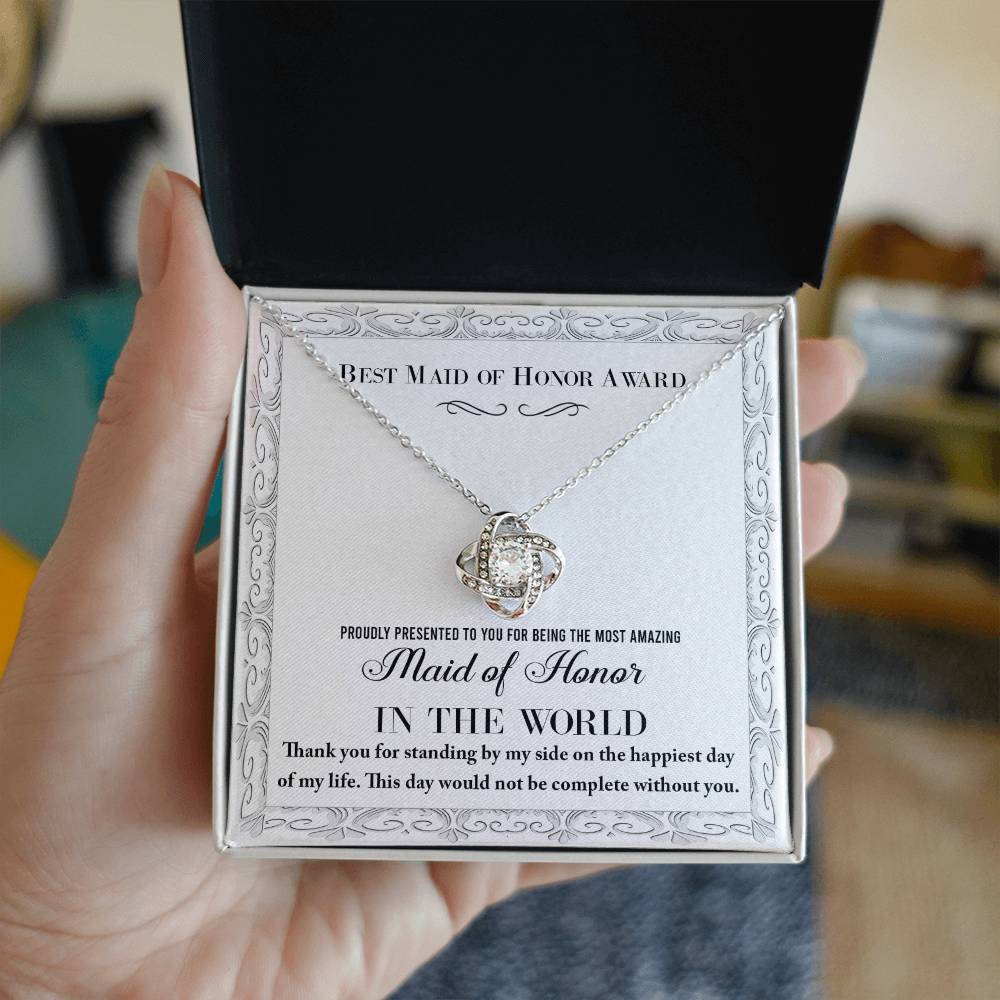 A "To Maid of Honor, Of My Life - Love Knot Necklace," crafted in 14k white gold with a heart-shaped pendant adorned with cubic zirconia crystals, sits elegantly in a jewelry box. The text reads, "Best Maid of Honor Award. Proudly presented to you for being the most amazing Maid of Honor in the world.