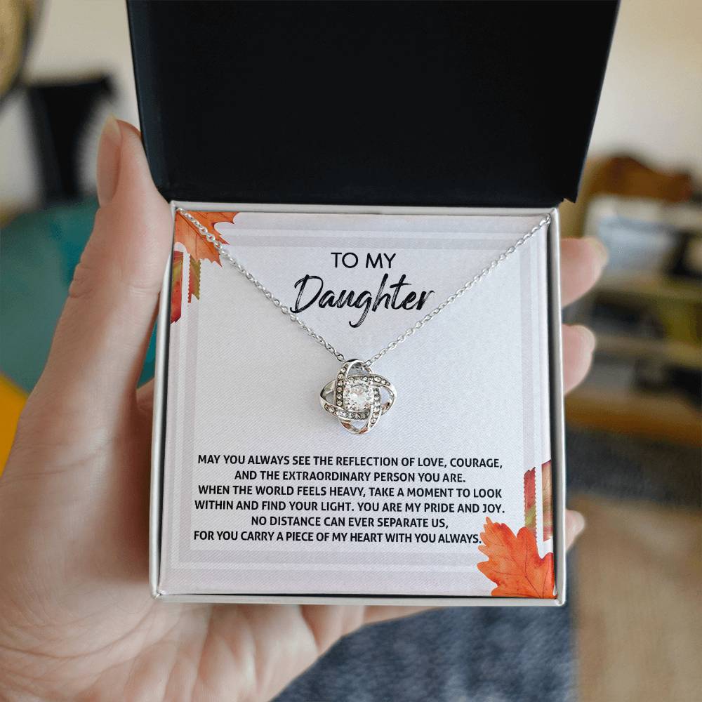 A person holds a Daughter-Find Your Light - Love Knot Necklace in a box with a message reading, "To my daughter... You are my pride and joy. No distance can ever separate us." The necklace, adorned with cubic zirconia crystals, is available in either a white gold or yellow gold finish.