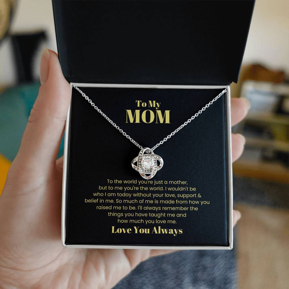 A hand holds an open box containing a stunning "To My Mom, To The World You Are Just A Mother - Love Knot Necklace" adorned with cubic zirconia crystals. The box lid has an inscribed message to "Mom," expressing gratitude and love, making it the perfect personalized gift.