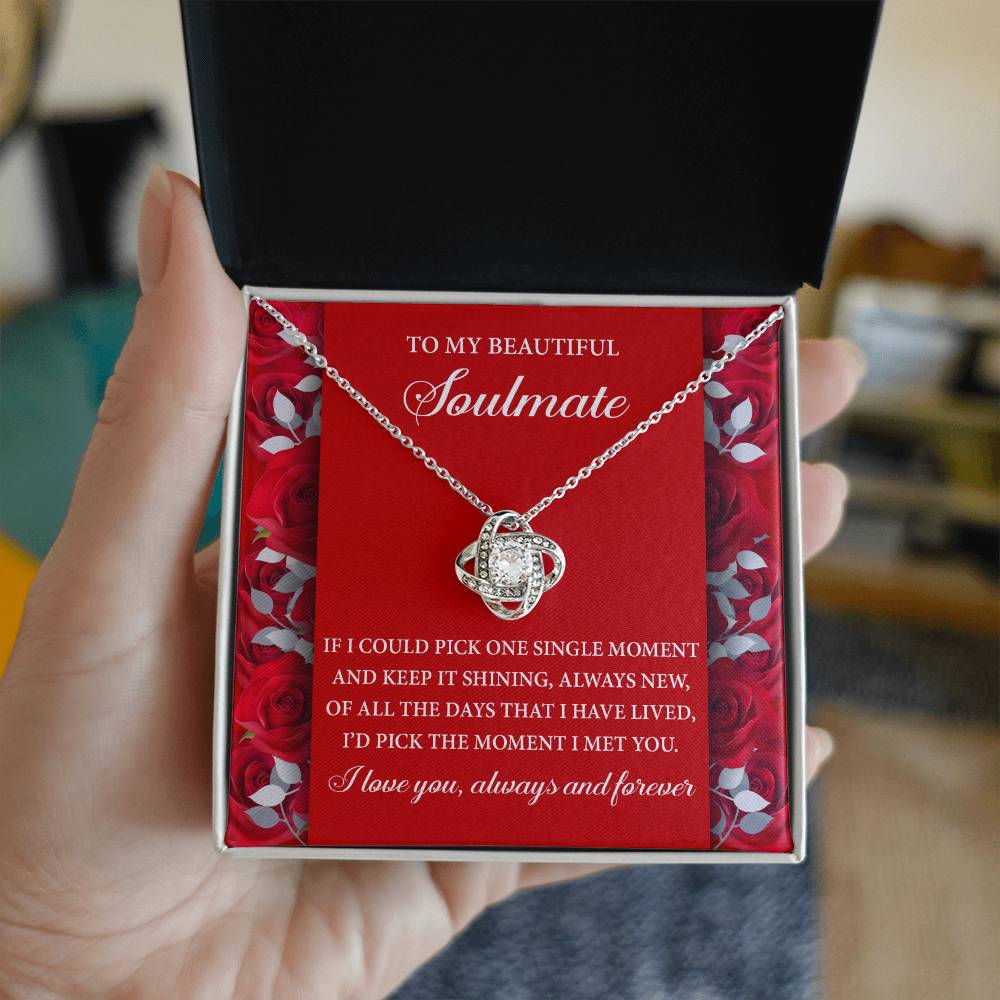 A hand holds a box with the "Soulmate-Keep It Shining - Love Knot Necklace," featuring a gold finish and cubic zirconia, above a card for a "beautiful soulmate," surrounded by roses.