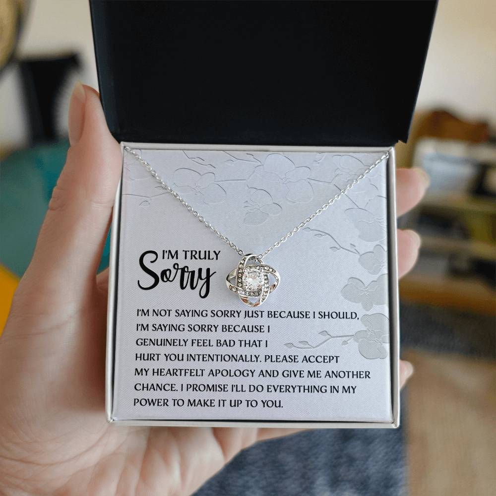 Introducing the Sorry-Feel Bad - Love Knot Necklace, featuring a pendant on a card with an apology message: "I'm truly sorry. I'm not saying sorry just because I should..." and continues with more heartfelt text. The necklace showcases a sparkling cubic zirconia crystal set in your choice of white gold or yellow gold finish, all against a light floral design background.