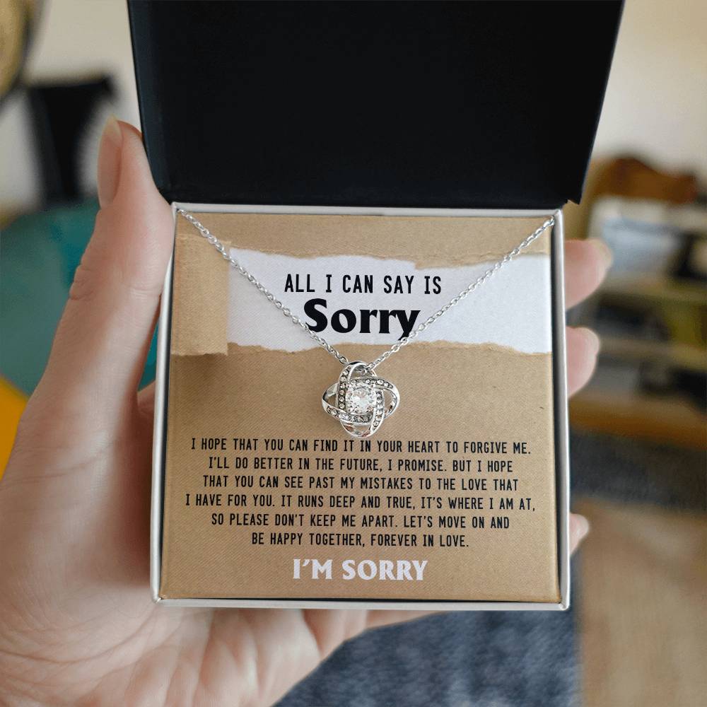 A hand holds an open jewelry box featuring a Sorry-Keep Me Apart - Love Knot Necklace adorned with cubic zirconia crystals. The box contains an apology note that reads: “All I can say is sorry. I hope that you can find it in your heart to forgive me…”.