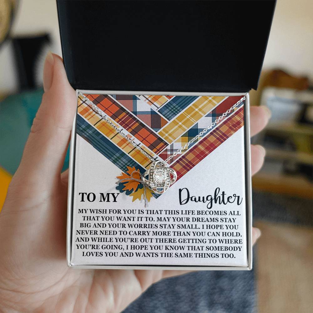 A hand holds an open box with the "Daughter-The Same Things - Love Knot Necklace," adorned with cubic zirconia crystals and a message inside. The message reads, "To My Daughter..." followed by a heartfelt wish for her life. The background features a plaid pattern and autumn leaves.