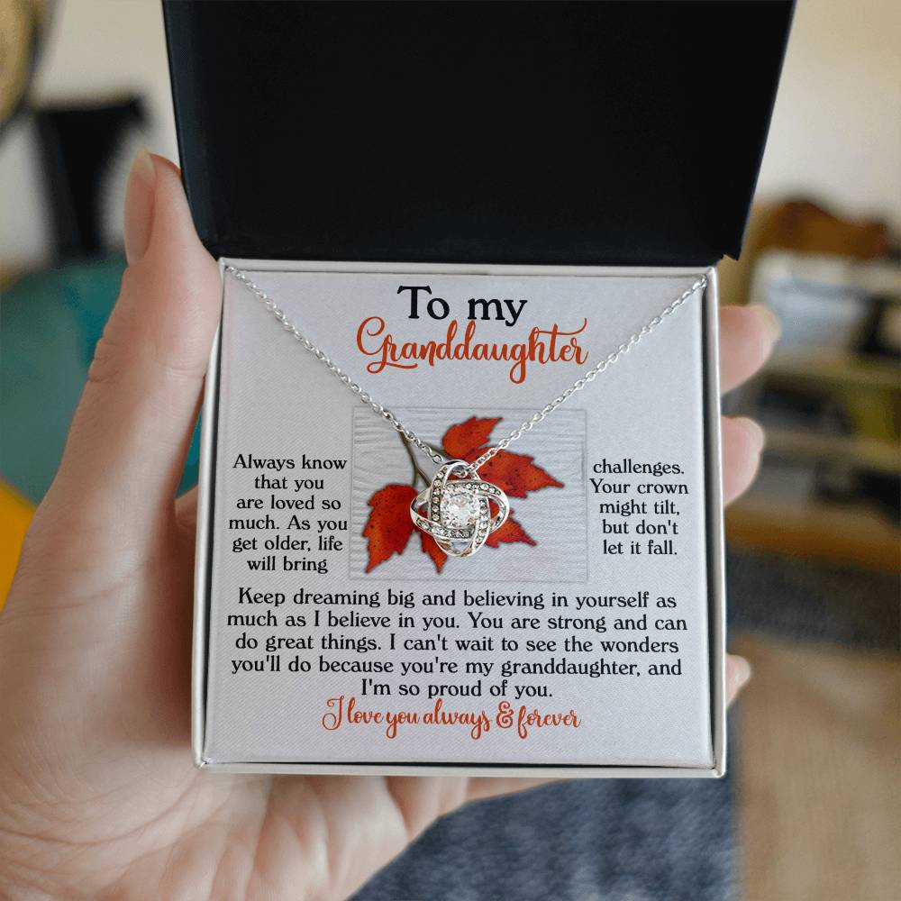 A hand holds an open box revealing the "Granddaughter-Do Great Things" Love Knot Necklace adorned with cubic zirconia. The box features an autumn leaf design and contains a heartfelt message for a granddaughter.