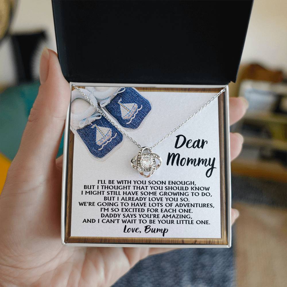 A hand holds an open jewelry box containing the "To Mom To Be, You're Amazing - Love Knot Necklace" adorned with cubic zirconia crystals, accompanied by a note addressed to "Mommy" from "Bump," expressing excitement for their upcoming adventures together.
