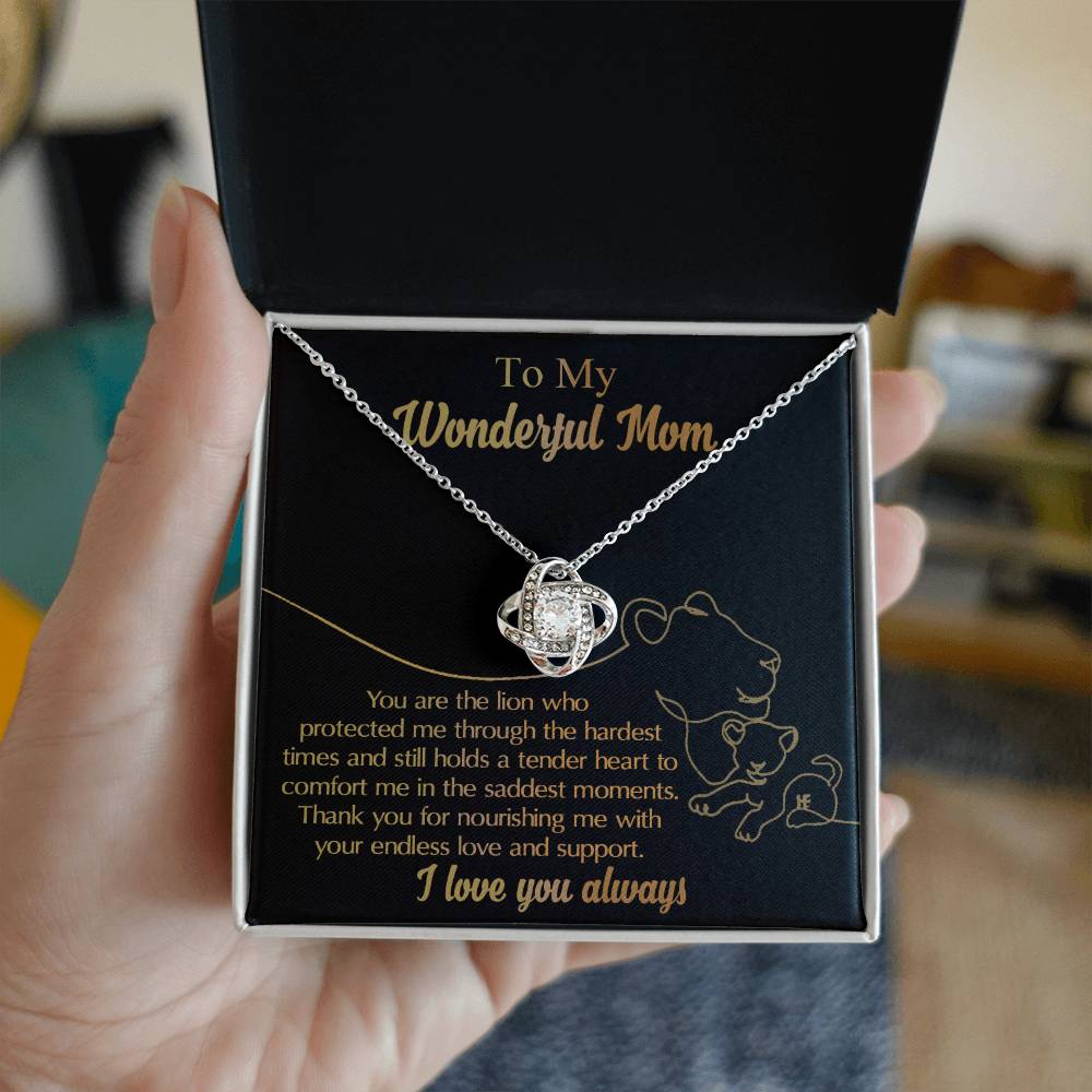 A hand holds an open gift box containing the "To Mom, The Lion - Love Knot Necklace," made of white gold and encrusted with cubic zirconia. Inside, a heartfelt message is printed, addressed "To My Wonderful Mom," expressing gratitude and love.