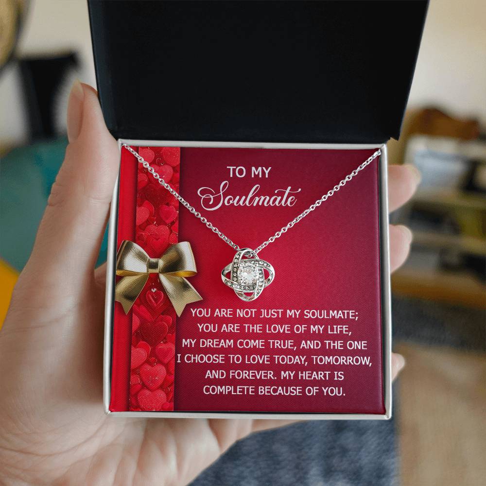 A person holds a jewelry box with the Soulmate-Because Of You - Love Knot Necklace, featuring cubic zirconia crystals against a red background and accompanied by a heartfelt soulmate message.