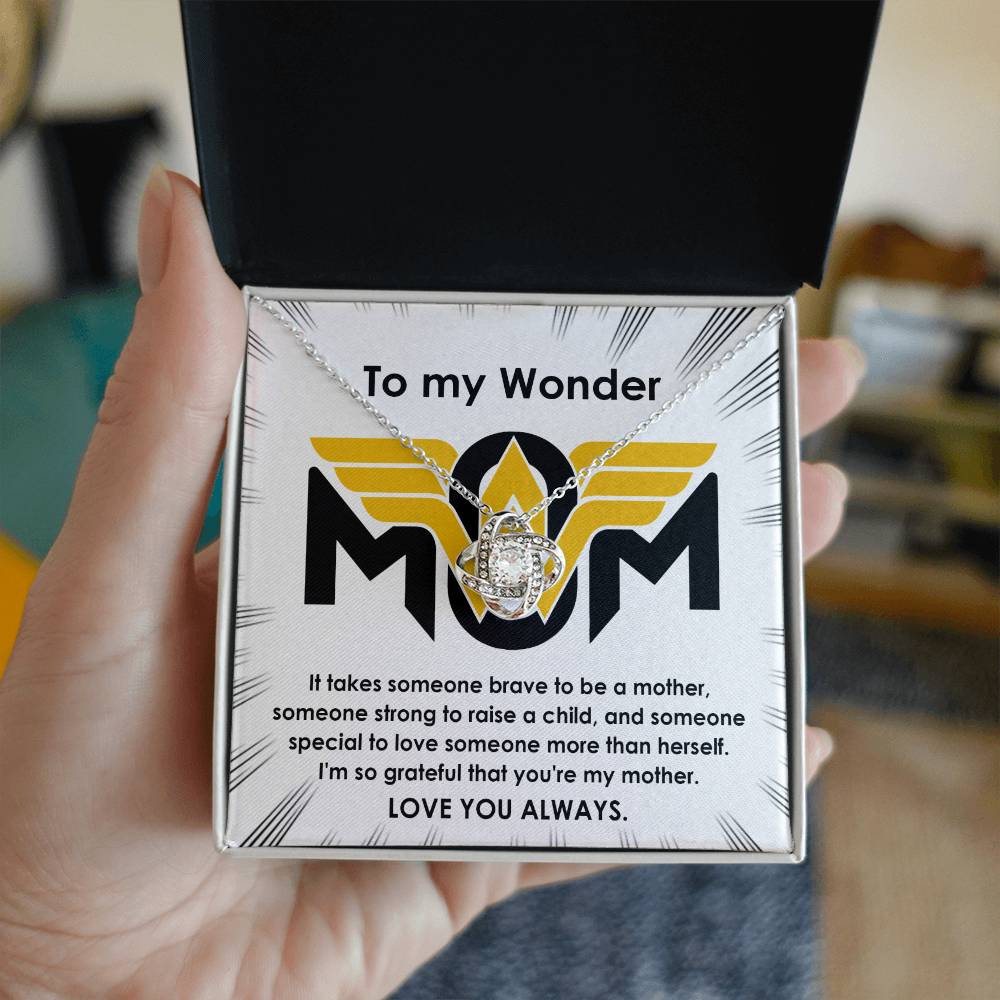 A hand holding an open jewelry box with a "To Mom, More Than Herself - Love Knot Necklace" adorned with cubic zirconia crystals. The box reads, "To my Wonder Mom," followed by a heartfelt message praising the recipient's strength and love.