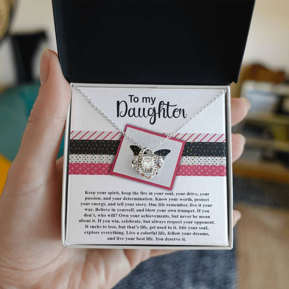 A hand holds an open jewelry box containing the "Daughter-Keep The Fire - Love Knot Necklace," which features intertwined rings and a cubic zirconia centerpiece. Addressed "To my Daughter," the necklace gleams with a gold finish. The background shows a room with a blurred-out object.