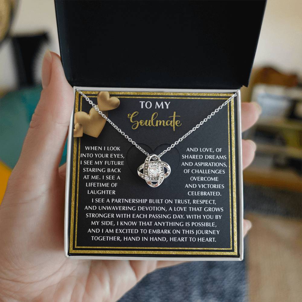 A person holds an open jewelry box revealing the Soulmate-Heart To Heart - Love Knot Necklace with cubic zirconia in a white gold finish. Inside the lid, a heartfelt message to a soulmate is beautifully decorated with two small golden hearts.
