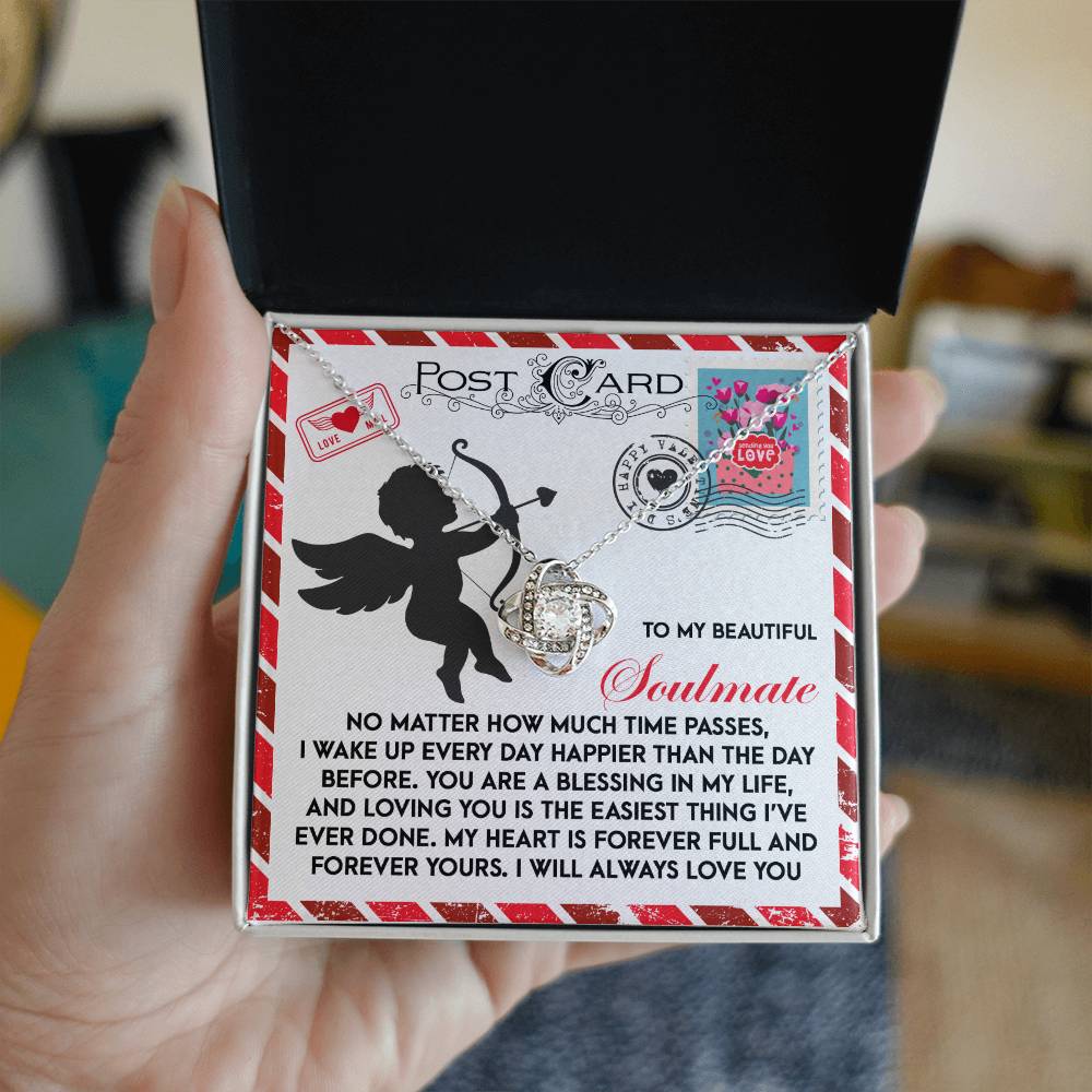 A person holds a gift box containing the "Soulmate-Forever Yours" Love Knot Necklace, adorned with sparkling cubic zirconia crystals. The box includes a card featuring a cupid design and a heartfelt message about cherishing your soulmate.