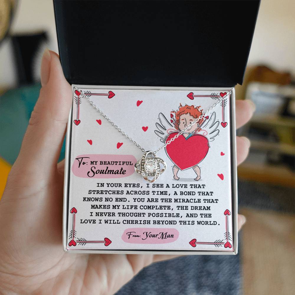 A hand holds an open gift box revealing the Soulmate-No End Love Knot Necklace in white gold with sparkling cubic zirconia. Behind it is a card featuring a heart and angel illustration with a printed love message for a soulmate.