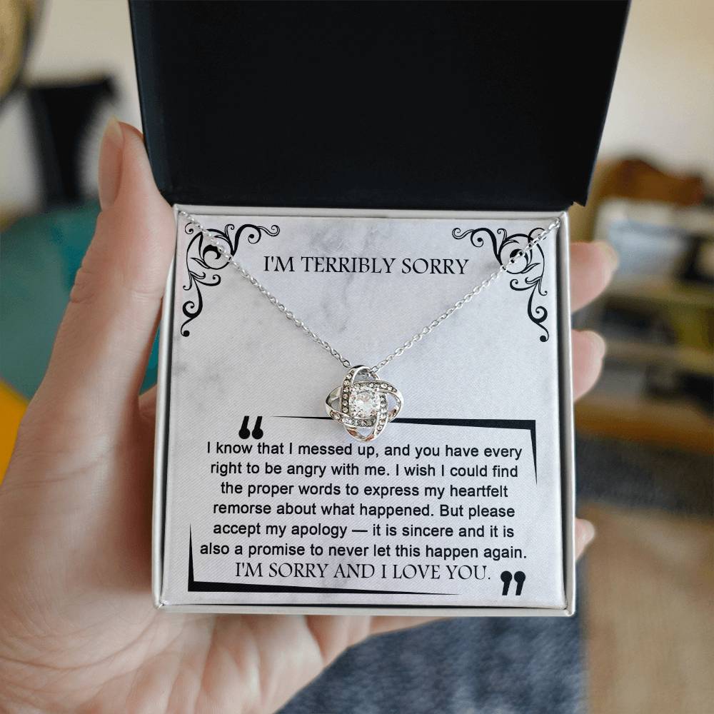 A hand holds an open box with the Sorry-Angry With Me - Love Knot Necklace, adorned with premium cubic zirconia crystals, accompanied by a heartfelt apology message that reads, "I know that I messed up, and you have every right to be angry with me... I'M SORRY AND I LOVE YOU.