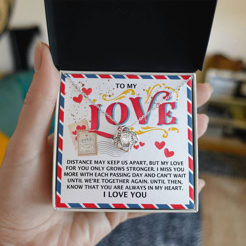 A person holds an open box, unveiling the radiant "Soulmate-In My Heart - Love Knot Necklace" alongside a heartfelt message, "TO MY LOVE," expressing how much they are missed and the enduring power of love across distances.