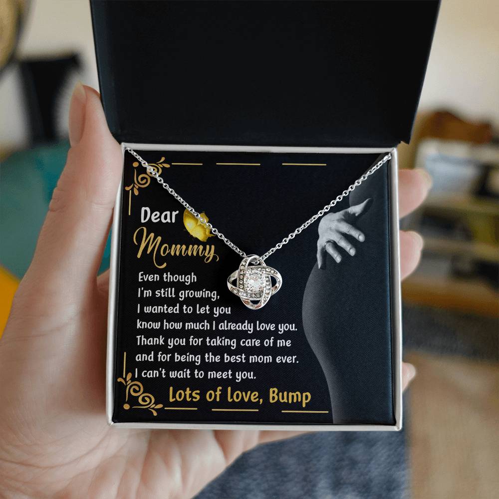A hand holds an open jewelry box displaying the "To Mama To Be, Best Mom Ever - Love Knot Necklace" adorned with cubic zirconia crystals. The box is lined with a printed message addressed to "Mommy" from "Bump," expressing love and appreciation.