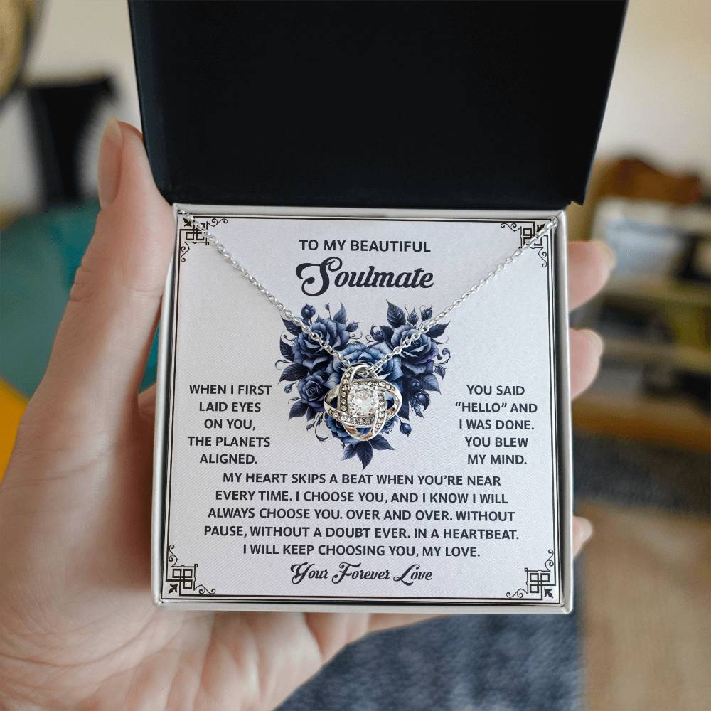 A hand holds an open jewelry box, displaying the Soulmate-Skips A Beat - Love Knot Necklace with cubic zirconia crystals. Inside, a romantic message to a soulmate makes it the perfect personalized gift.