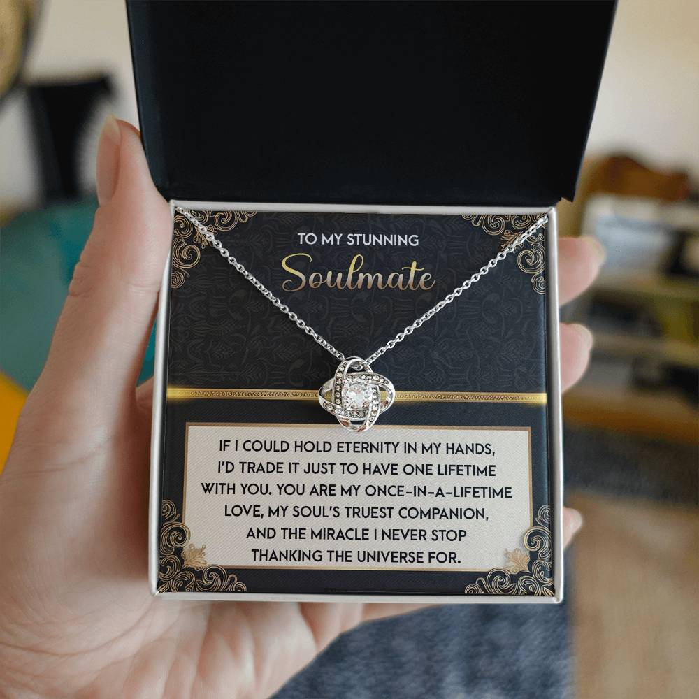 A hand holds an open jewelry box with the "Soulmate-Truest Companion - Love Knot Necklace," elegantly crafted in white gold finish and adorned with sparkling cubic zirconia. The lid reads "Soulmate" alongside a heartfelt message.