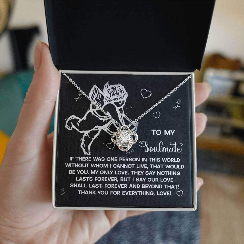A hand presents an open jewelry box unveiling the Soulmate-Lasts Forever Love Knot Necklace. It features intertwined hearts with cubic zirconia accents, available in white or yellow gold finishes. The inside lid is decorated with an angel illustration and a romantic message.