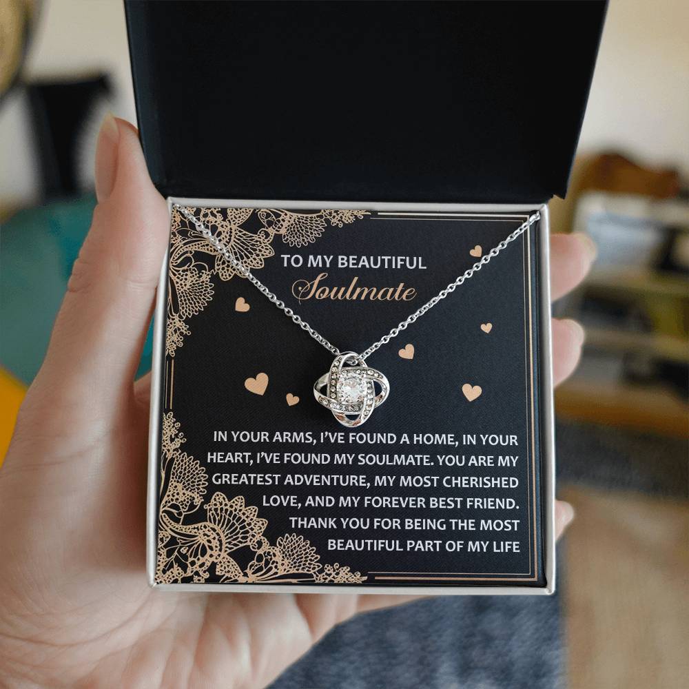 A person holds an open box showcasing the Soulmate-Beautiful Part - Love Knot Necklace, featuring a gold finish with sparkling cubic zirconia and a heartfelt message to their "beautiful soulmate" on a decorative card.