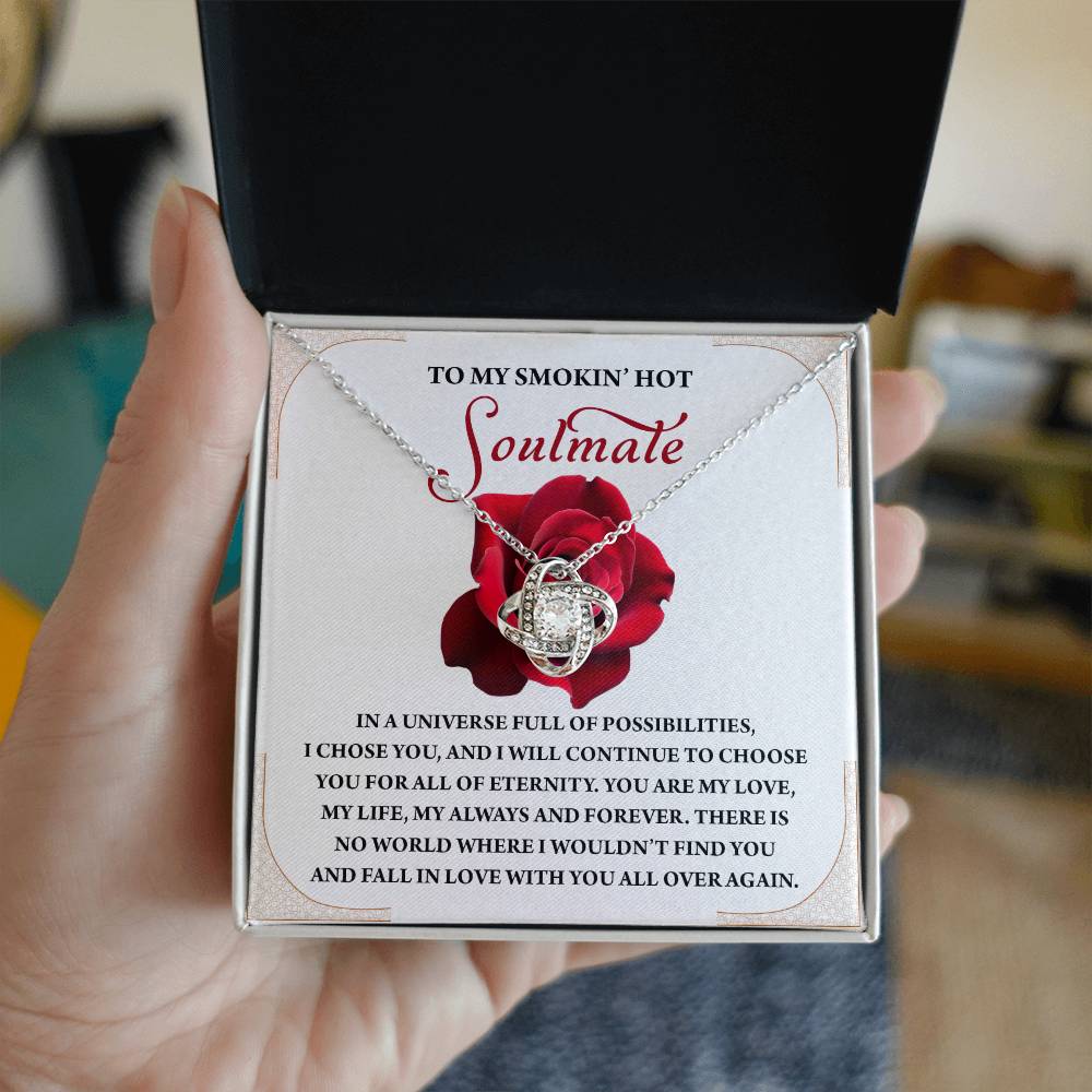 A hand presents an open jewelry box showcasing the Soulmate-Over Again - Love Knot Necklace, sparkling with cubic zirconia crystals, set against a card featuring a rose and a touching message to a soulmate.