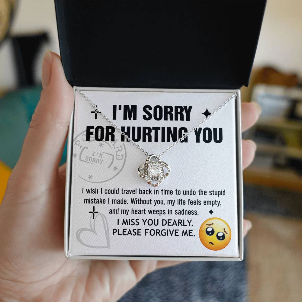 A Sorry-Travel Back - Love Knot Necklace with a white gold finish is displayed on a card featuring the text: "I'M SORRY FOR HURTING YOU," along with an apology note and a sad face emoji at the bottom right.