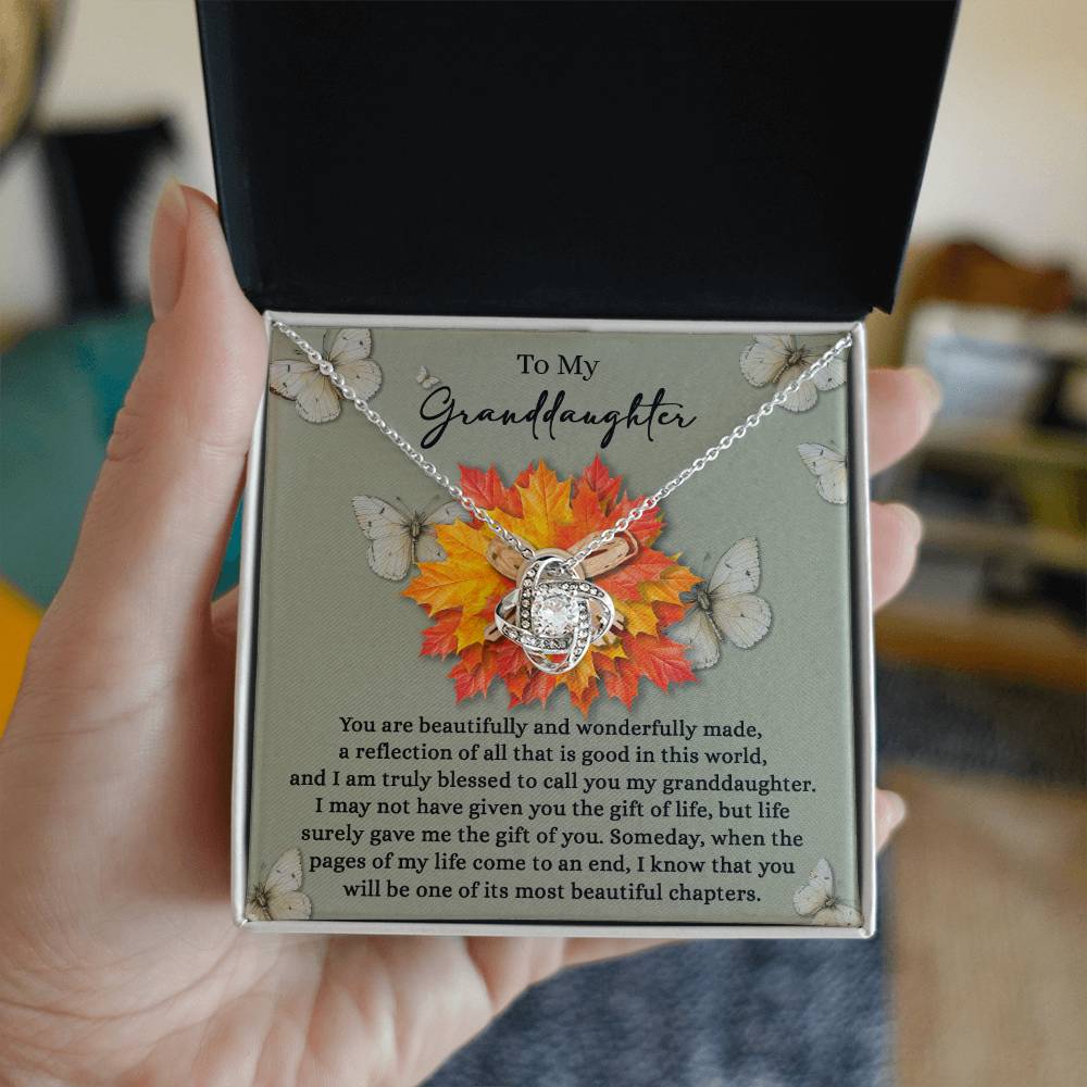 A person holds an open jewelry box featuring the "Granddaughter-Gift Of You - Love Knot Necklace," sparkling with cubic zirconia crystals. The box displays a heartfelt message for a granddaughter and is embellished with butterfly and autumn leaf designs.