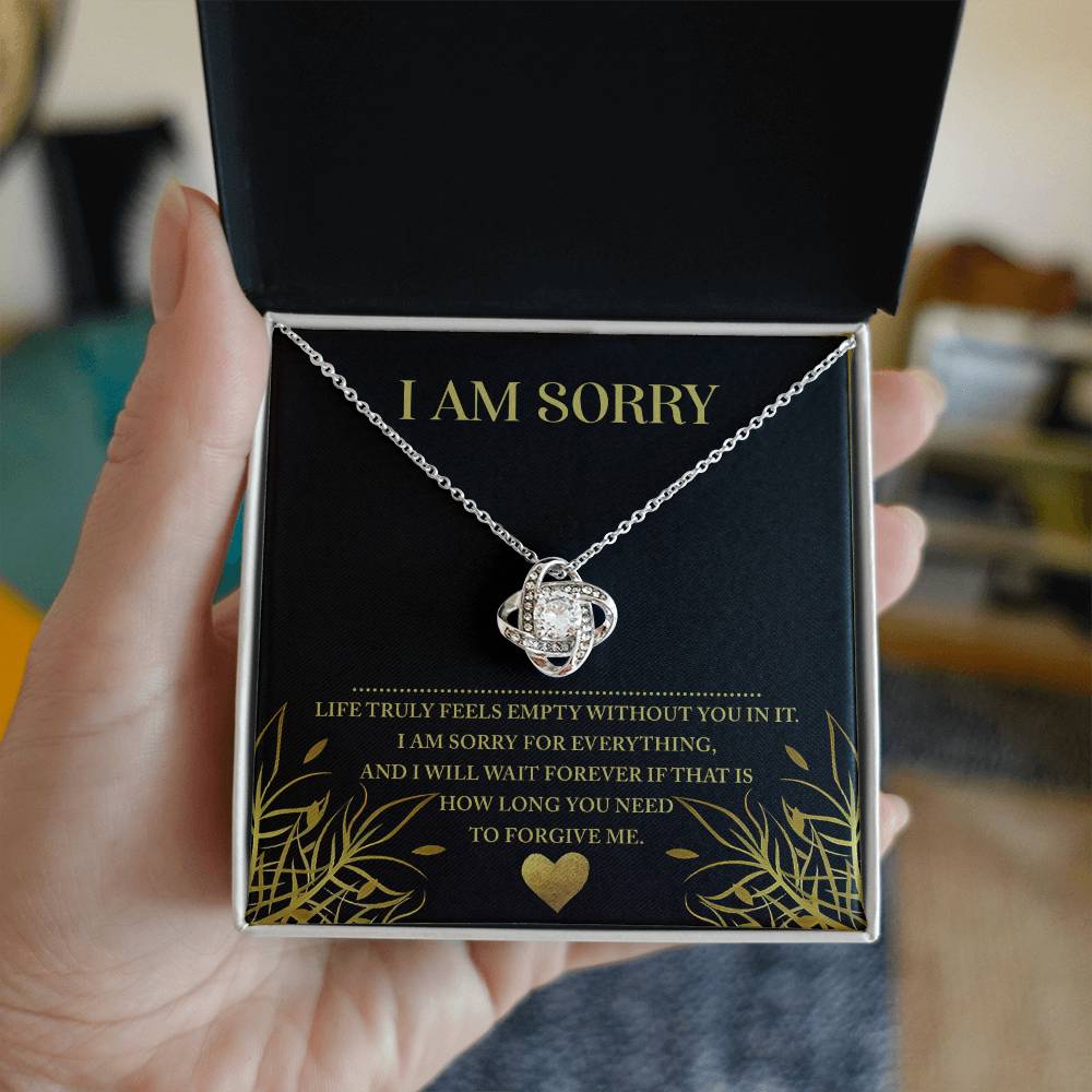 A hand holding an open jewelry box reveals a stunning Sorry-Without You - Love Knot Necklace adorned with cubic zirconia crystals and an apology message: "I am sorry. Life truly feels empty without you. I am sorry for everything, and I will wait forever if that is how long you need to forgive me.