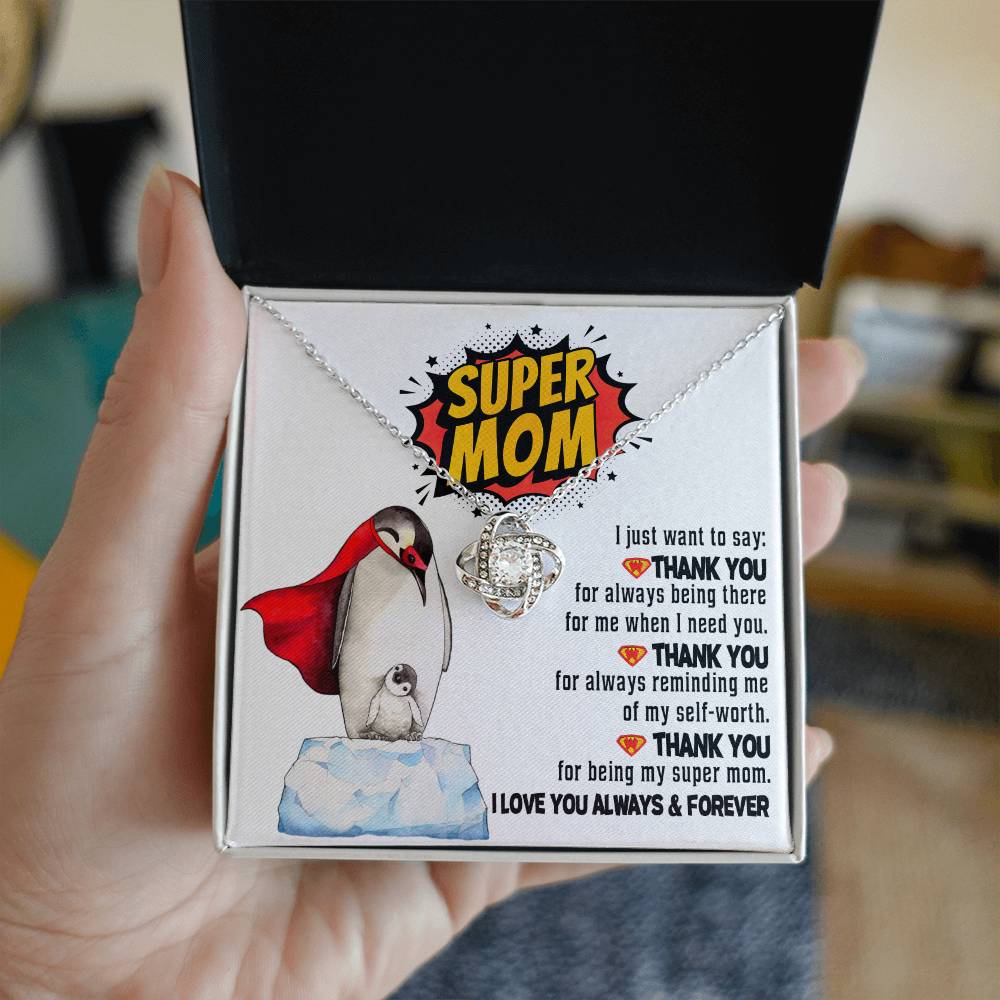 A hand holds an open jewelry box containing the "To Mom, Super Mom - Love Knot Necklace." The box lid reads "SUPER MOM" next to an image of a penguin and chick, accompanied by a thank-you message in appreciation of a mother's unbreakable bond.
