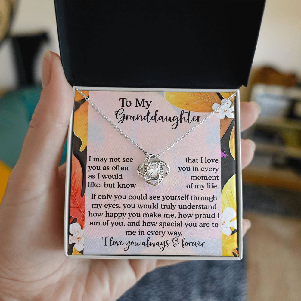 A hand presents an open box revealing the Granddaughter-In Every Way - Love Knot Necklace, which showcases a stunning pendant adorned with a brilliant cubic zirconia and set in a sophisticated white gold finish. Inside the box is a touching message dedicated to a granddaughter, accented by yellow and white flowers.